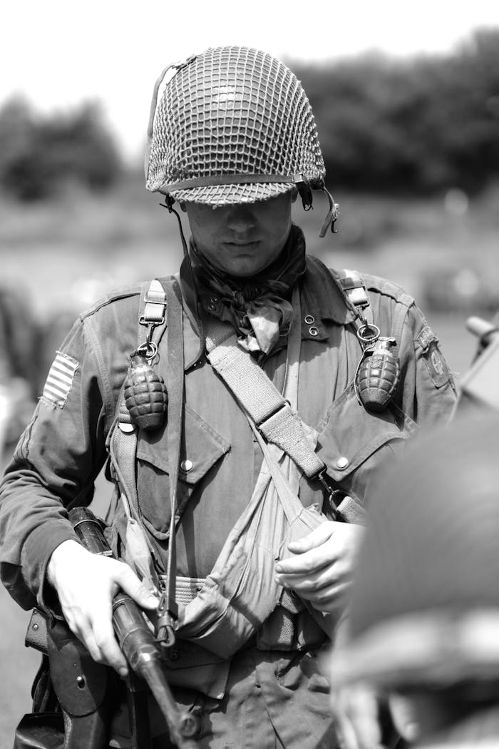 Black and white photo of a soldier in WWII-era uniform holding a rifle with grenades and helmet.